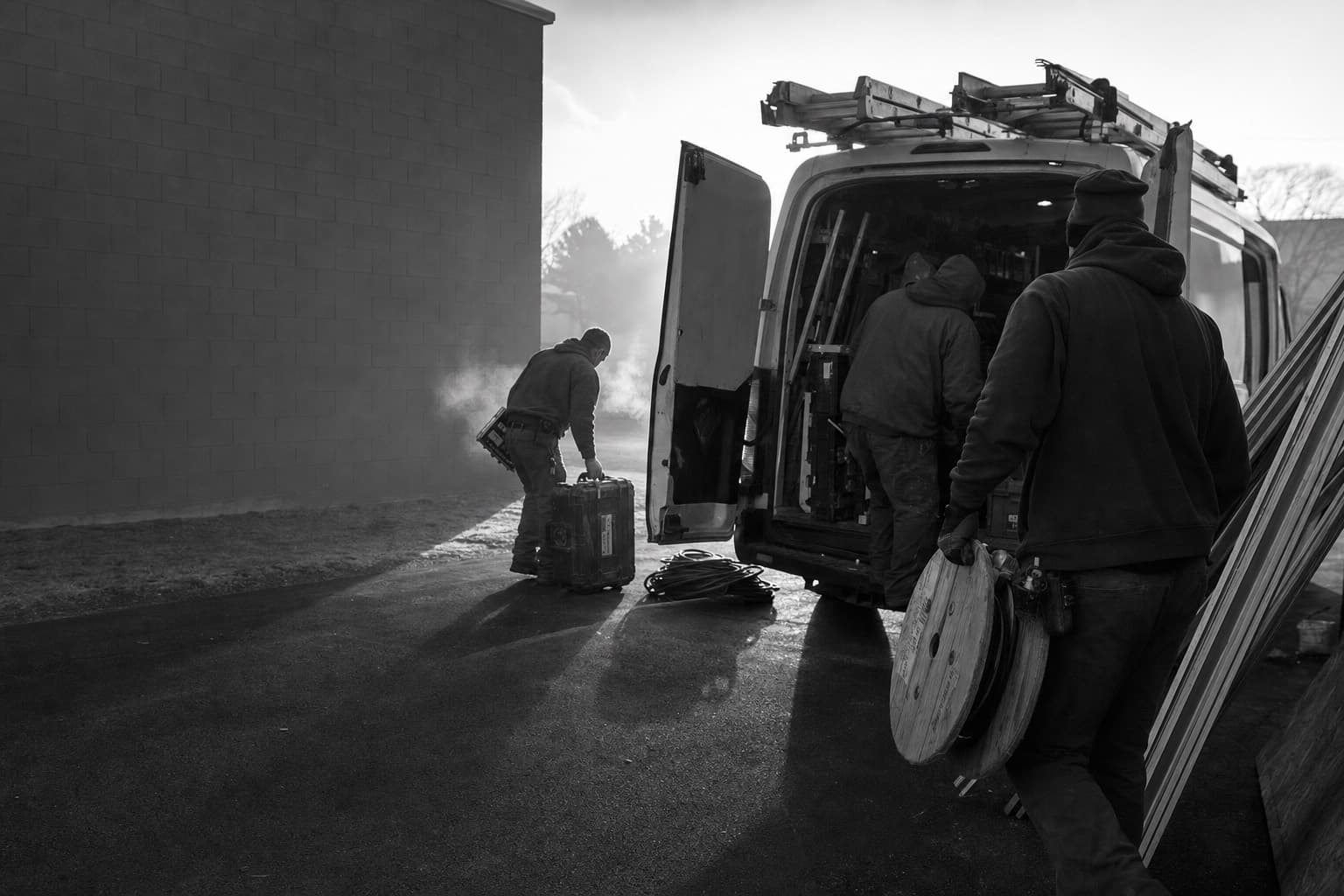 An electrical crew loads tools into a work van at dawn, breath visible in the cold.